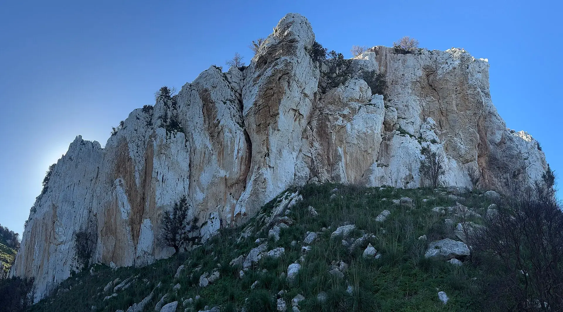 Parete di calcare chiaro con striature arancioni sopra un pendio erboso, vicino a Caltavuturo, sotto il cielo azzurro