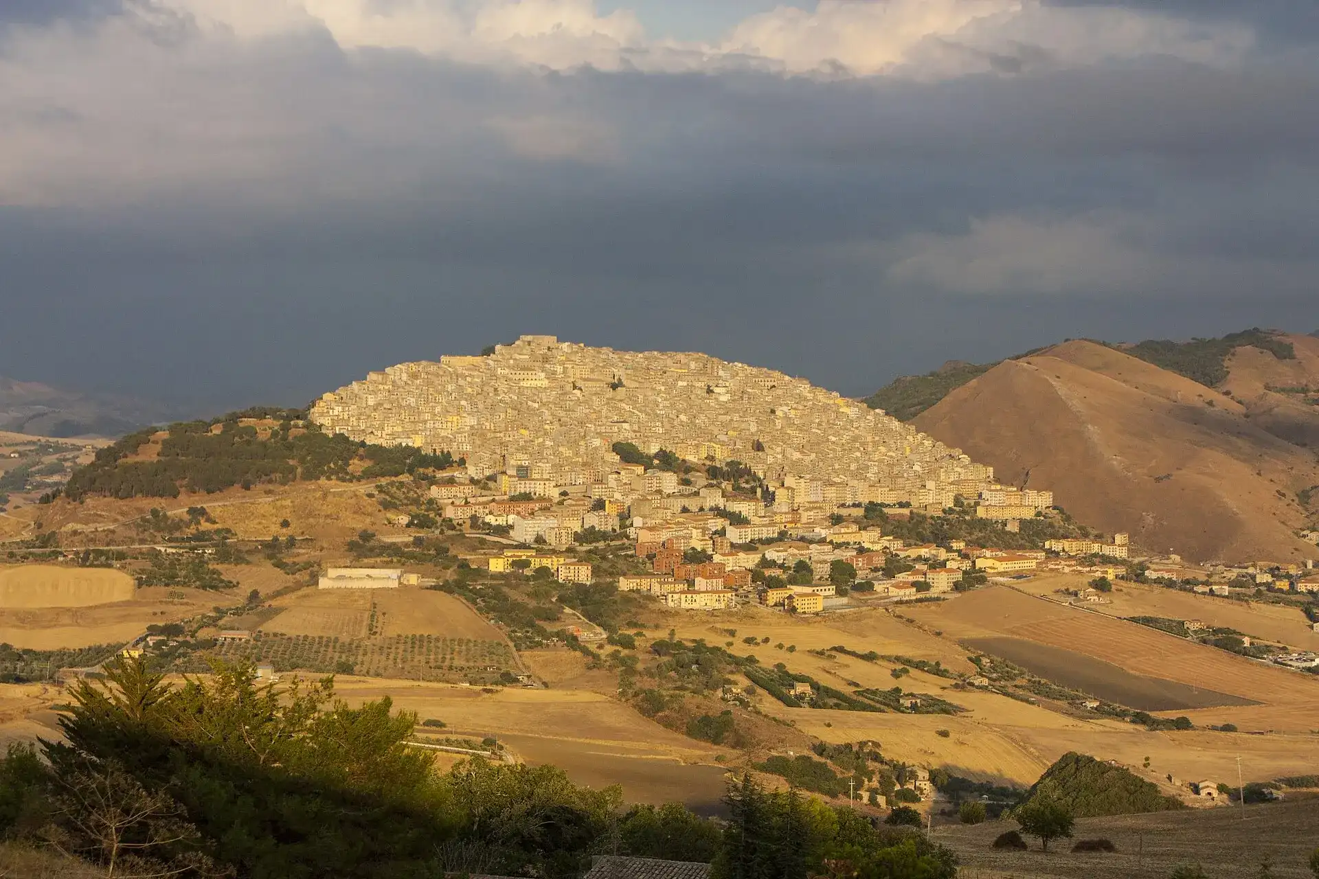 Gangi illuminato dal sole del pomeriggio sulla sua collina, tra campi di grano mietuto e un cielo di temporale