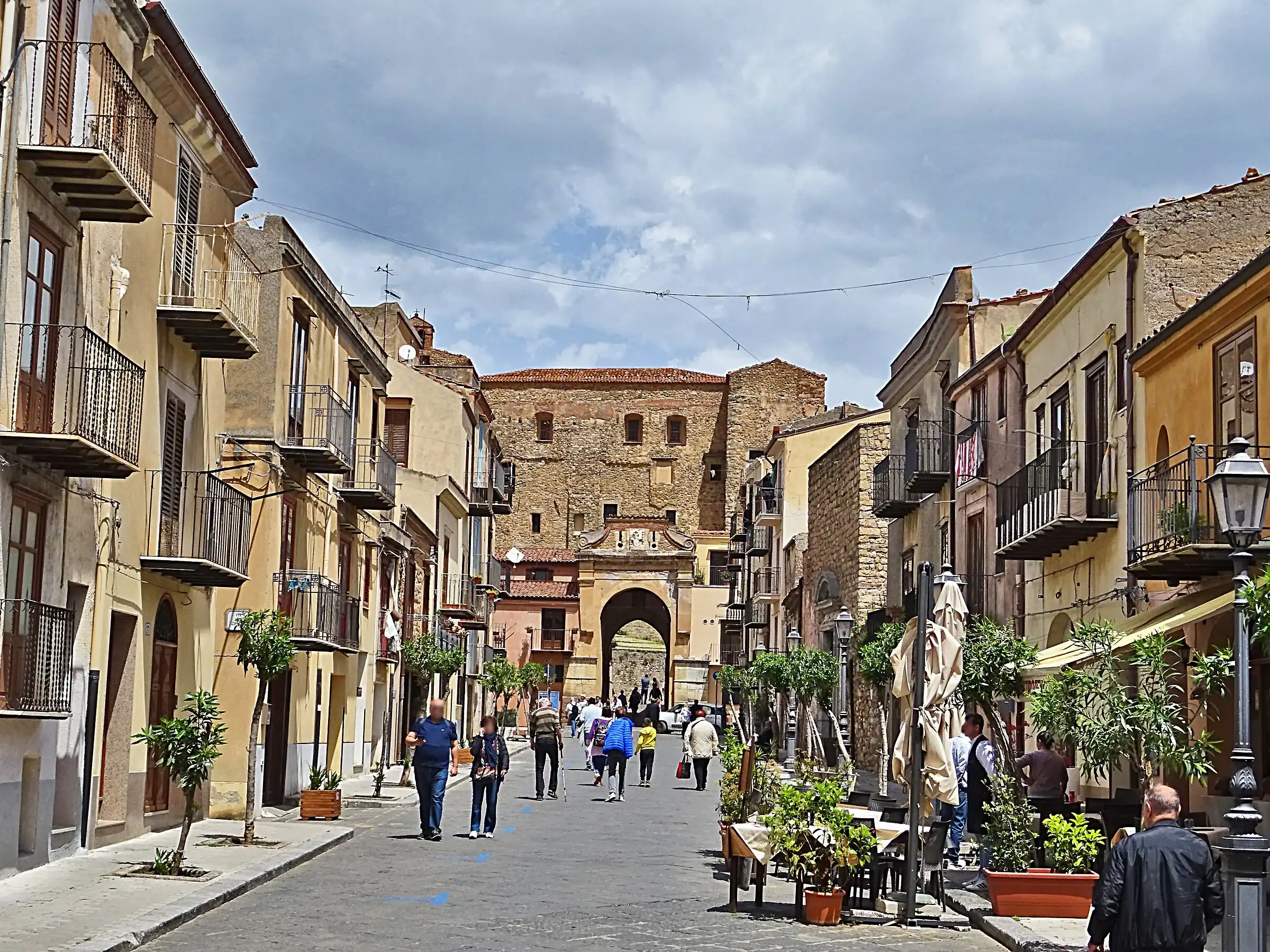 Via Sant’Anna a Castelbuono, con negozi, piante e passanti, che sale verso l’arco e il castello