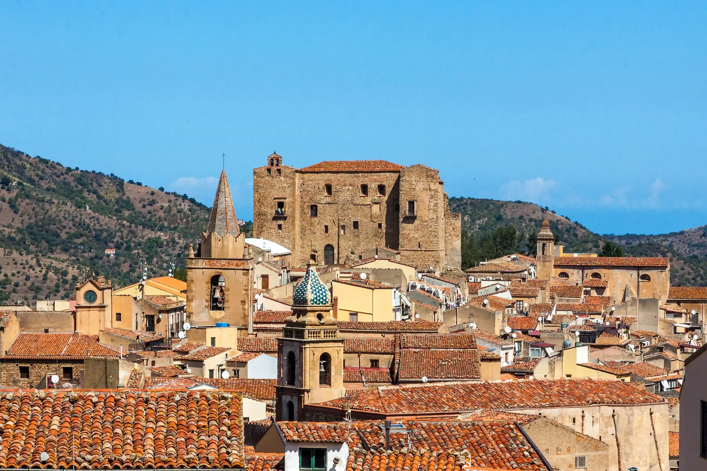 I tetti di Castelbuono con il castello, i campanili e una cupola di maioliche, tra le colline verdi
