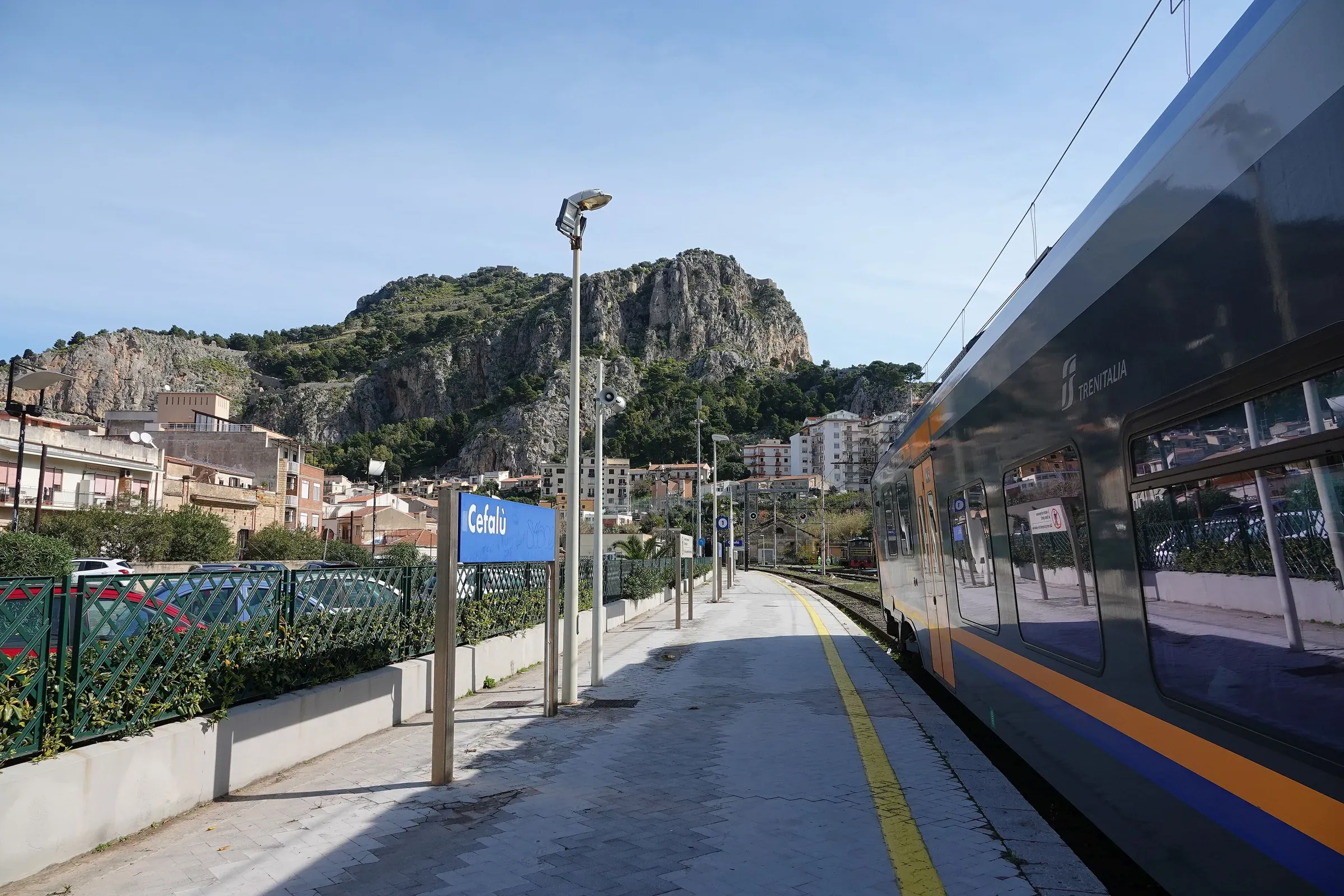 Un treno regionale di Trenitalia fermo alla stazione di Cefalù, con il cartello blu e la Rocca sullo sfondo