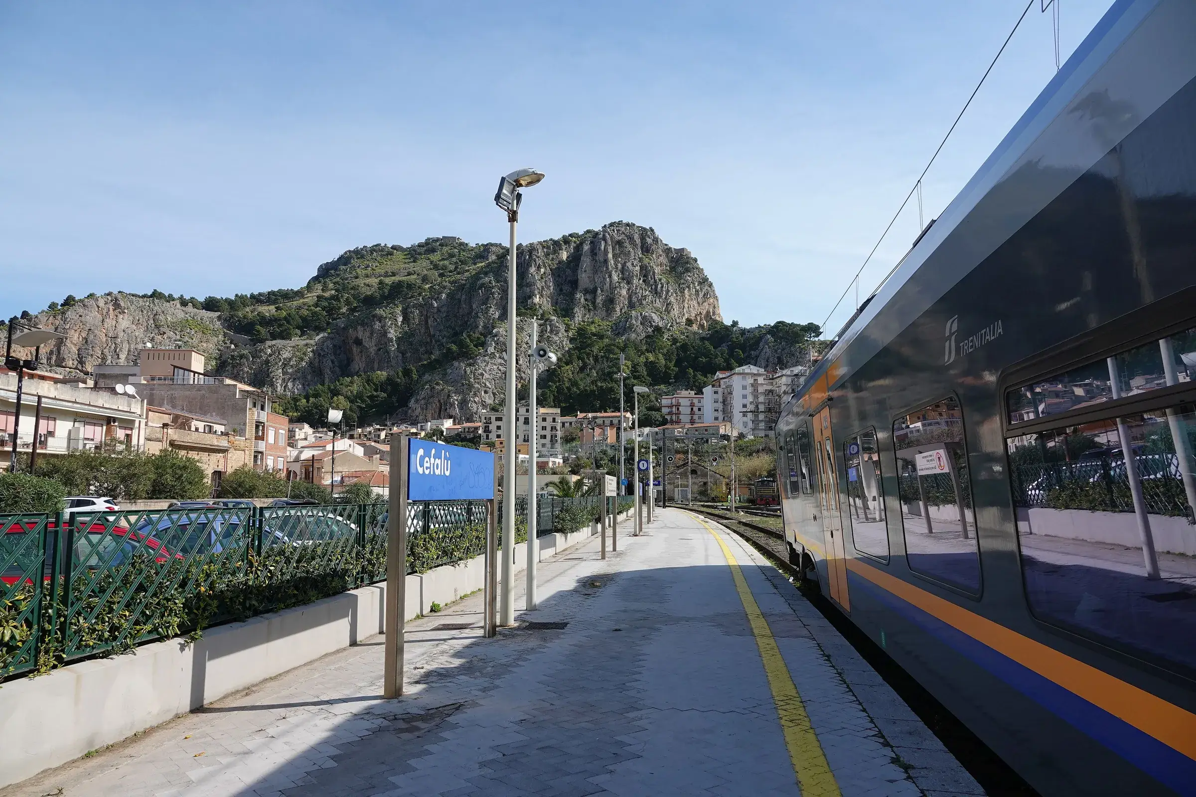 Un treno regionale di Trenitalia fermo alla stazione di Cefalù, con il cartello blu e la Rocca sullo sfondo