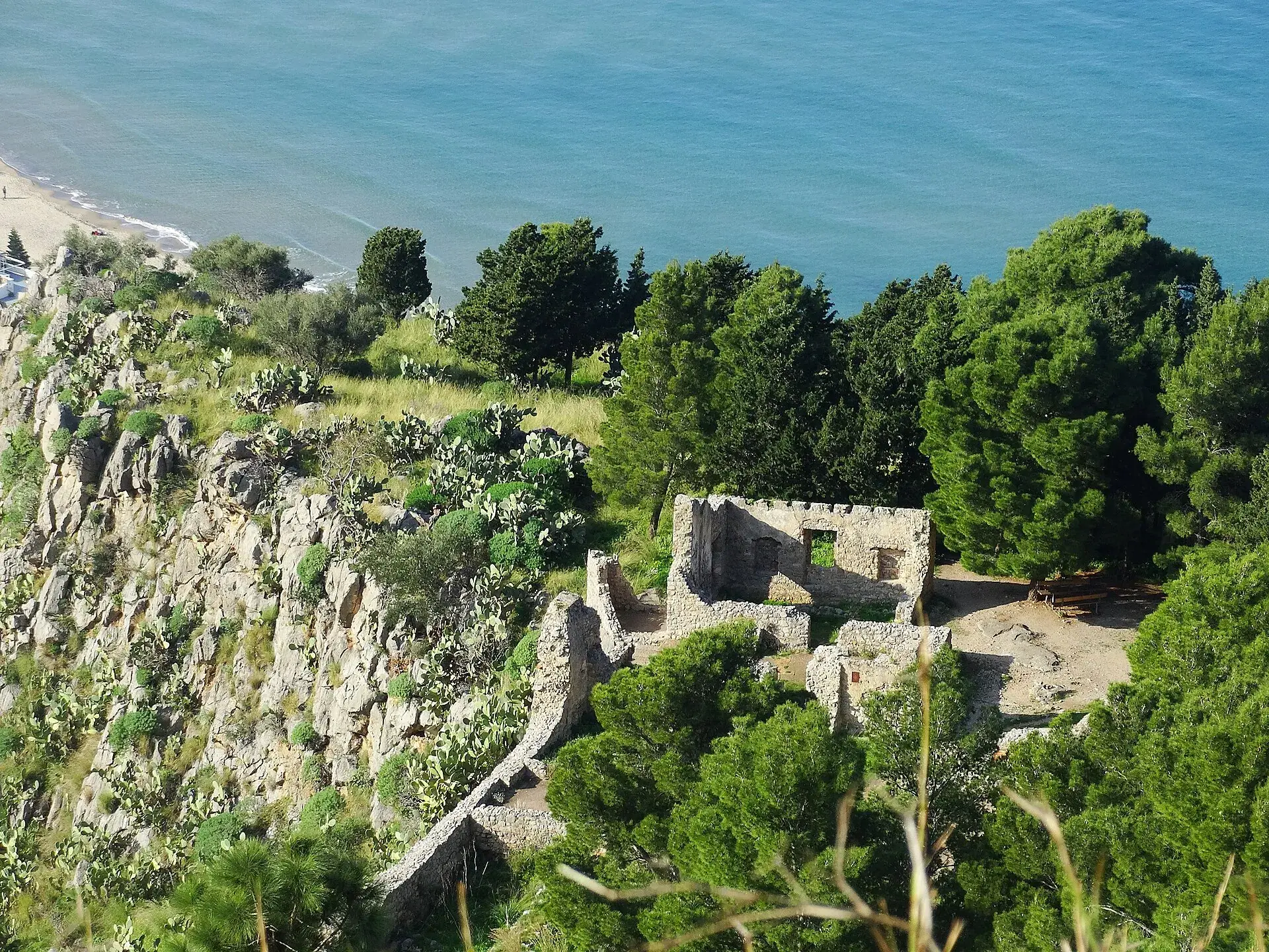 In cima alla Rocca di Cefalù i resti in pietra di un edificio tra i pini, con la spiaggia e il mare azzurro sotto