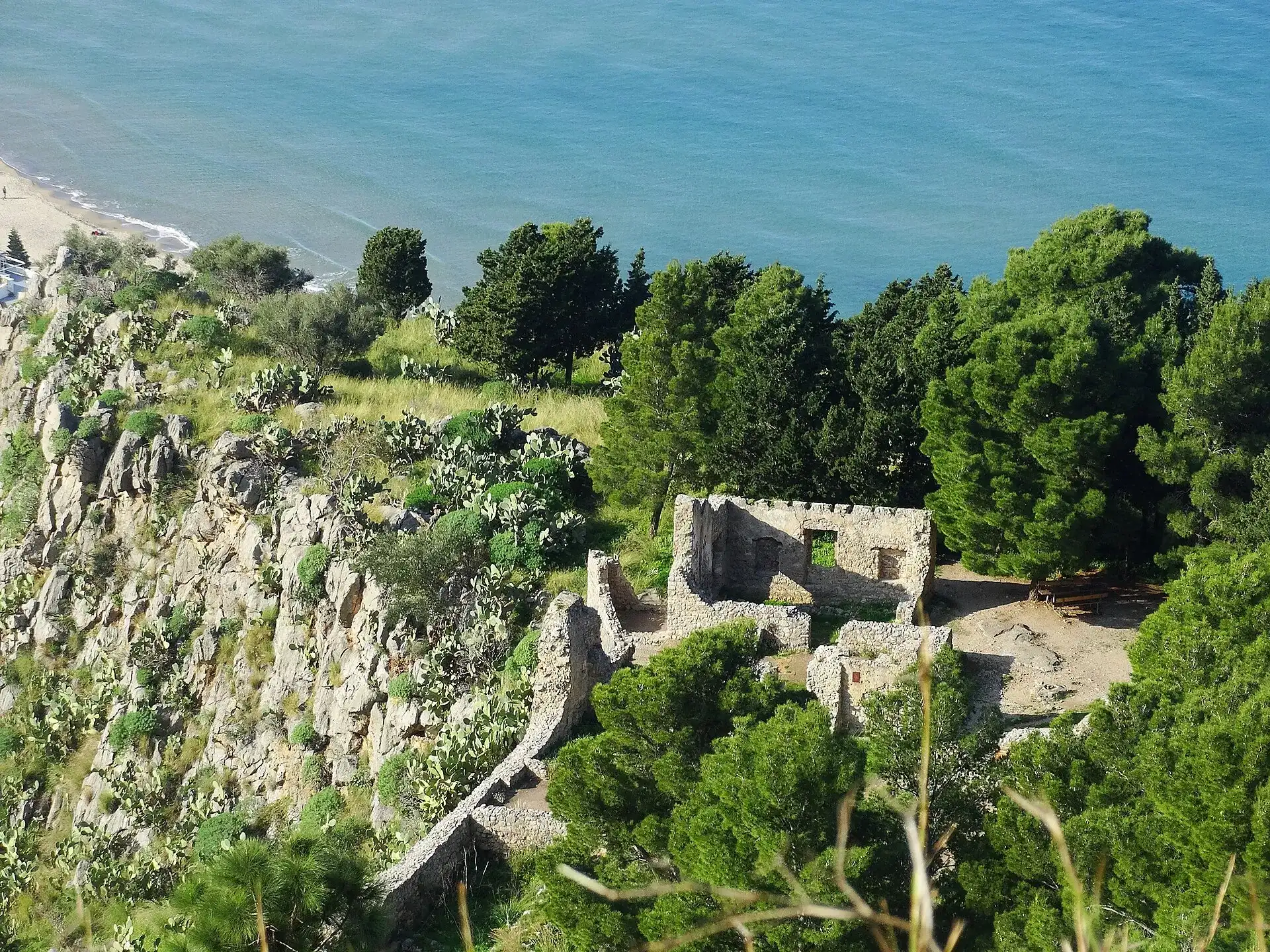 In cima alla Rocca di Cefalù i resti in pietra di un edificio tra i pini, con la spiaggia e il mare azzurro sotto