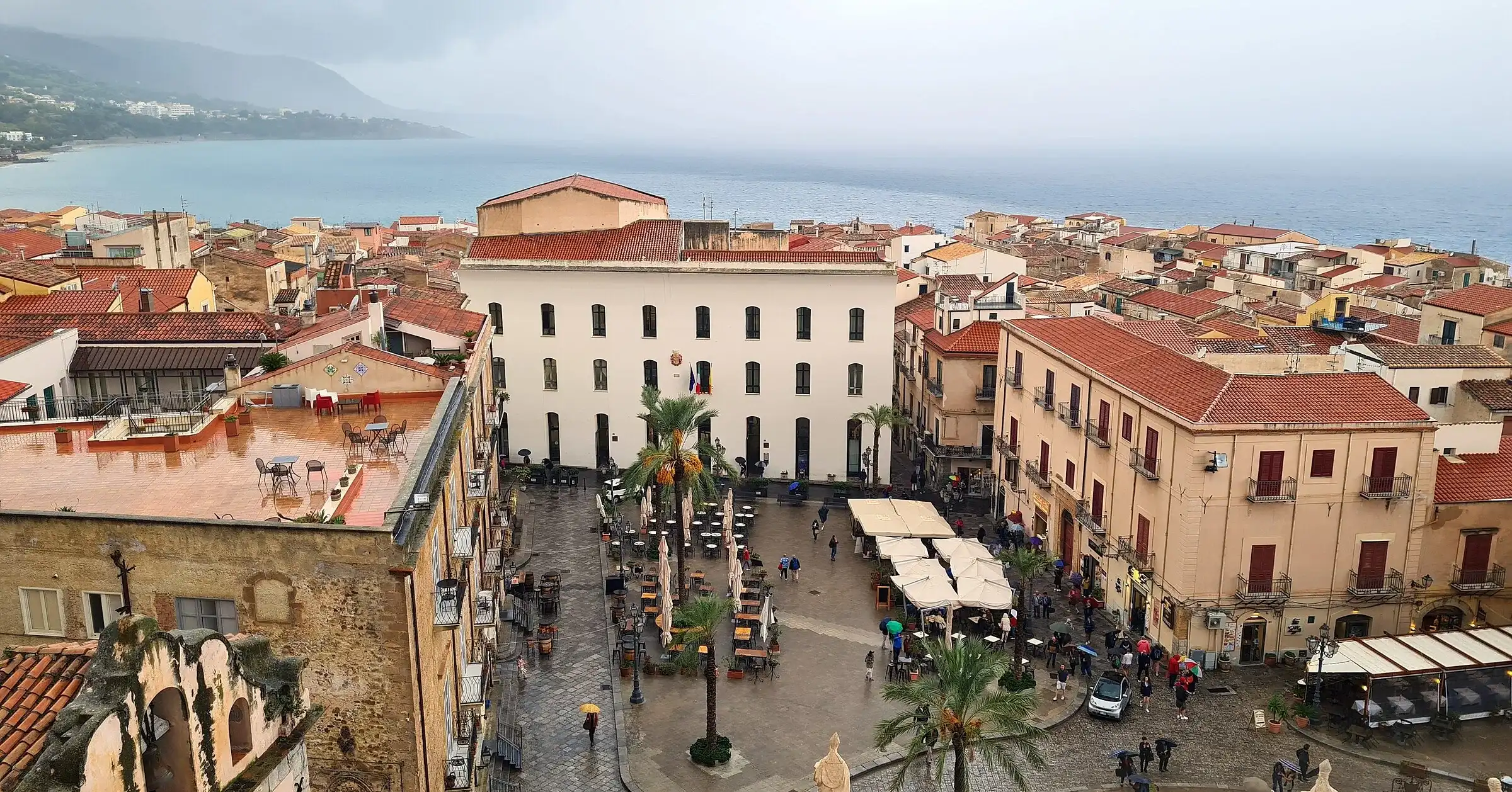 Piazza Duomo a Cefalù vista dall’alto, con i palazzi, i tavolini all’aperto e il mare sullo sfondo
