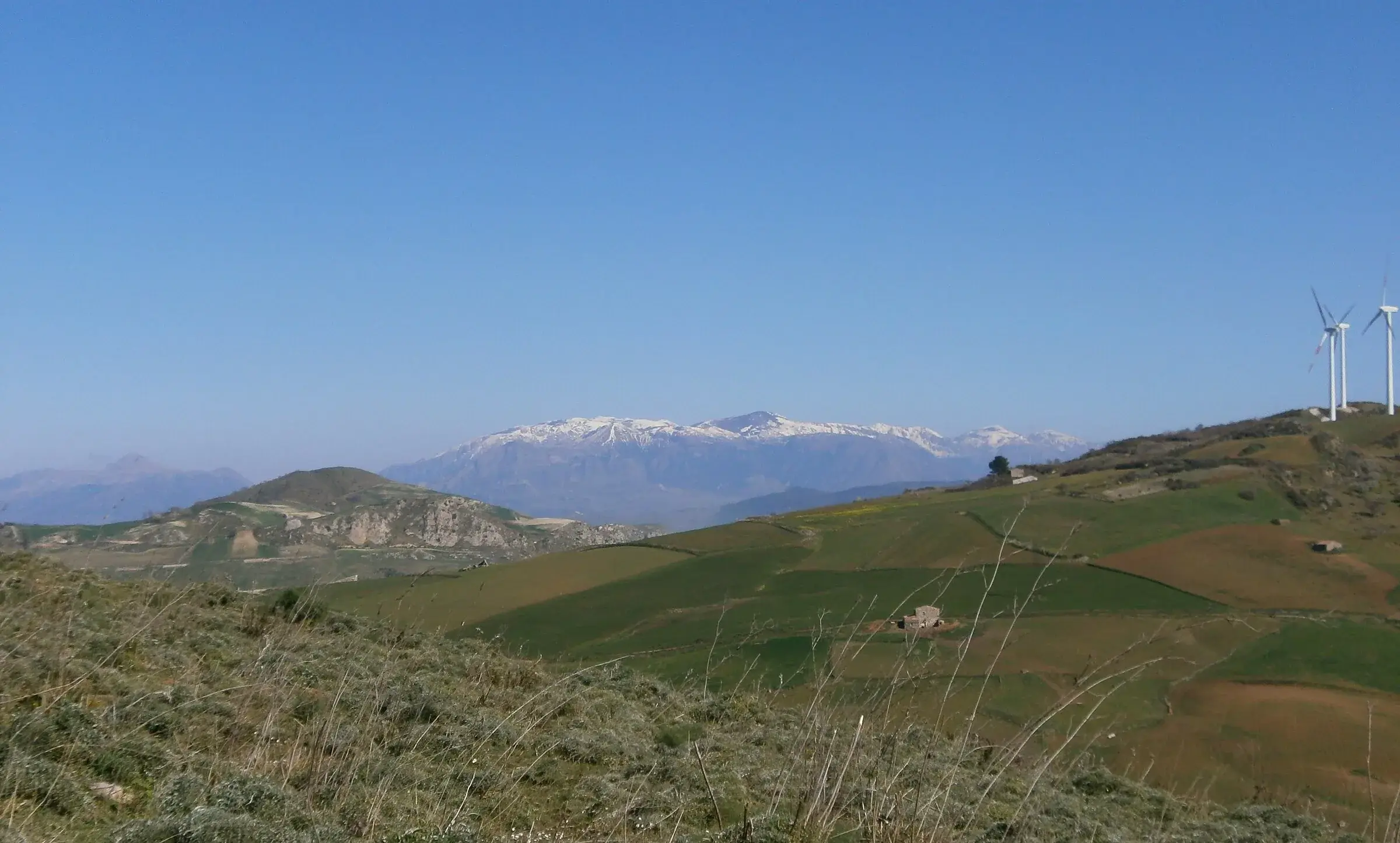 Le cime delle Madonie coperte di neve, viste da lontano oltre le colline coltivate, con due pale eoliche